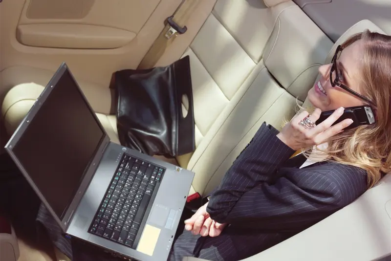 A businesswoman is talking on her smartphone and using a laptop while riding in a black car service, symbolizing Dallas Luxury Transportation.